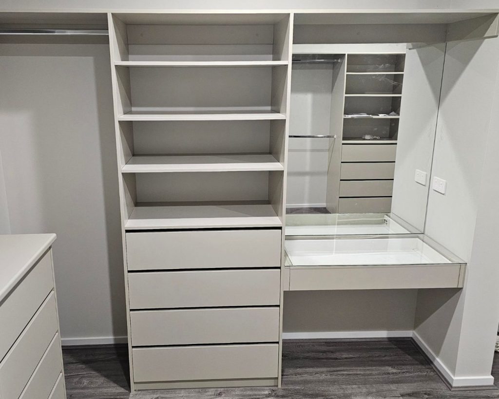 A built-in walk-in wardrobe interior with Laminex Paperbark finish featuring open shelving, a bank of drawers, a silver hanging rail, and a mirrored vanity shelf